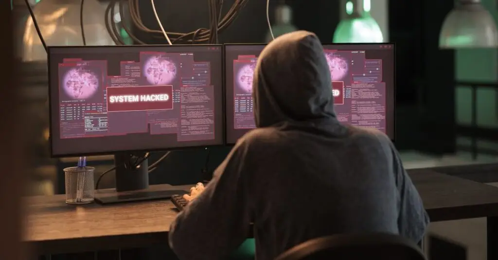 A hooded hacker accessing compromised computer systems displaying 'SYSTEM HACKED' warnings on dual monitors with code interfaces and data visualization, illustrating how macros pose a cybersecurity risk by granting attackers system-level access to sensitive files, security settings, and network resources.